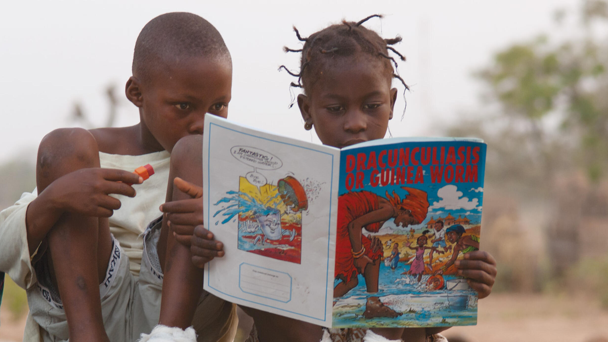 Two children crouch and read a comic book called "Dracunculiasis Or Guinea Worm" with a brightly illustrated cover.