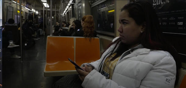 A person wearing a light-colored puffer coat sits in a subway car, holding a smartphone.