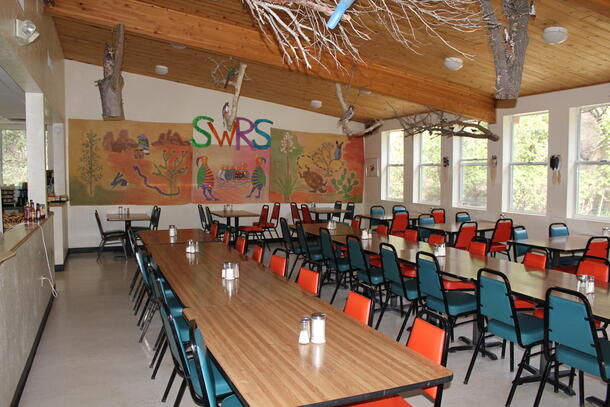 A dining hall with three long wooden tables, a few two-person tables in the back and a hand illustrated banner on the wall.