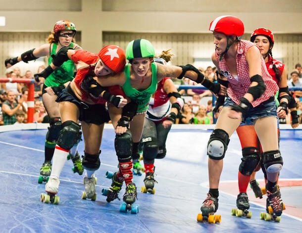 Six people in roller skates, helmets and knee pads skate in a circular roller derby rink, as one of them elbows another person out of the way.