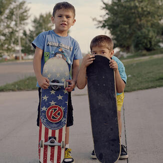 Two young boys stand with their skateboards on the sidewalk. One is crouching behind the board.
