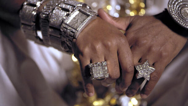 Two clasped hands covered in bling—a large ring on each hand and multiple, shining bracelets and watches on the wrists.