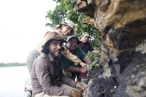 Three paleontologists and a local guide pose on the side of a rock formation, with a river visible behind them.