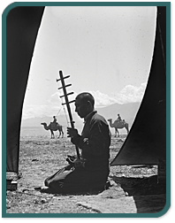 black and white photo of musician playing silk road stringed instrument with camels in the background