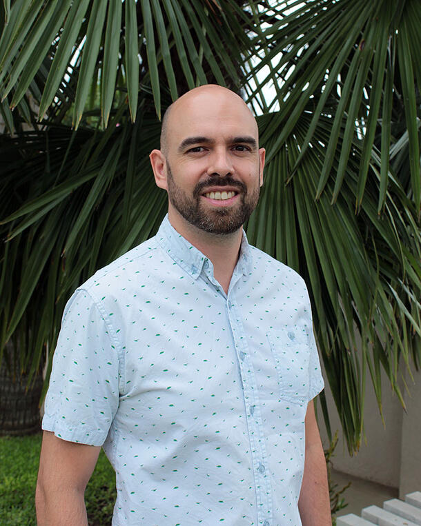 Edson Abreu poses for an outdoors headshot wearing a short-sleeved dress shirt, and standing in front of a palm tree.