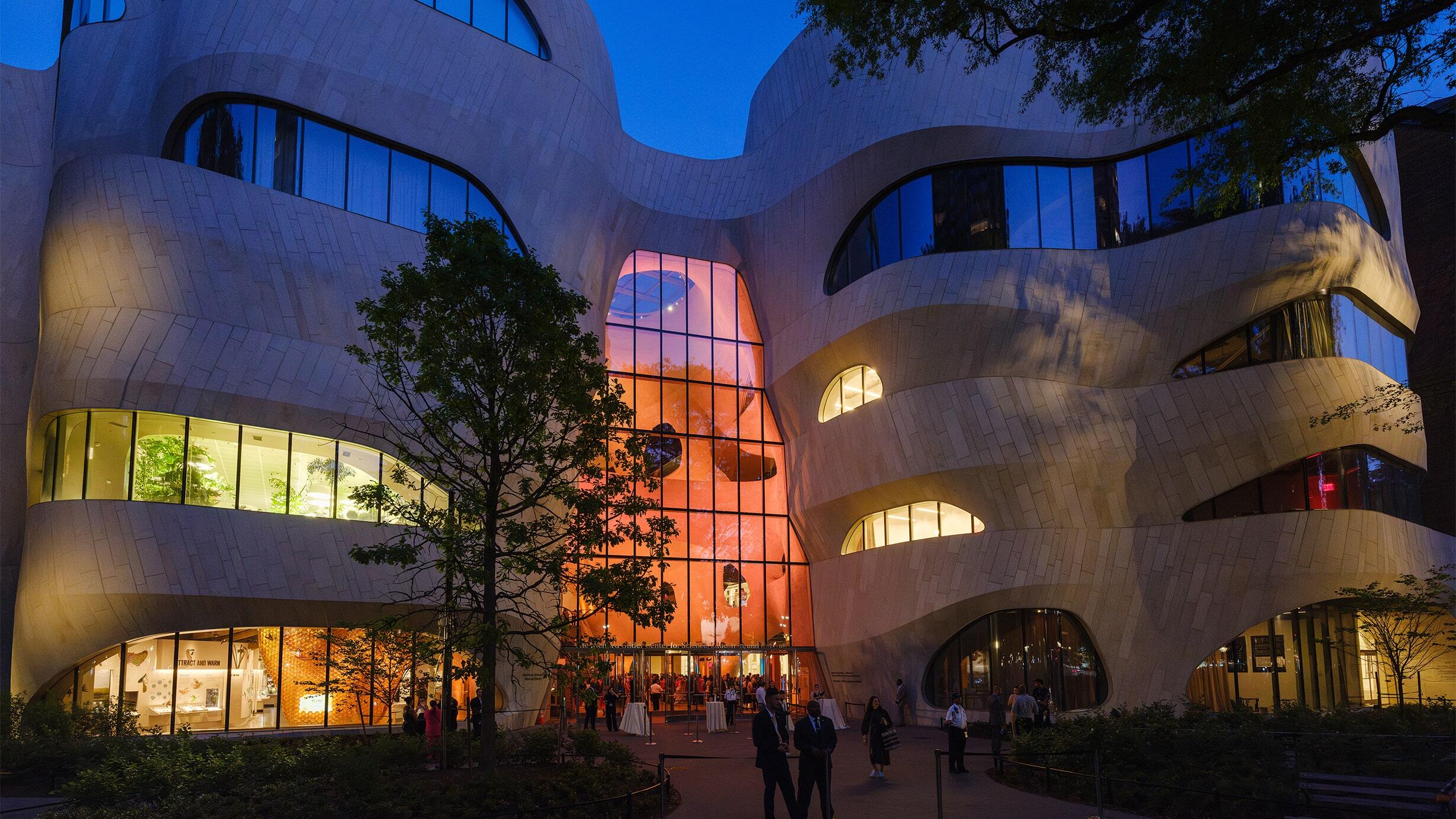 The Gilder Center at the American Museum of Natural History, seen at night. Bright lights emanate from within the curved exterior of the building.