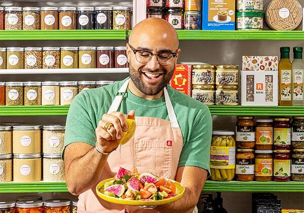 A photograph of chef Edy Massih, seen standing in front of shelves full of spices and dried goods, and squeezing fresh lemon onto a colorful salad.