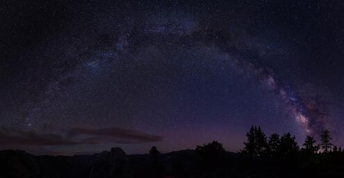 Trees and mountains under a wide view of the Milky Way arcing across the night sky. 